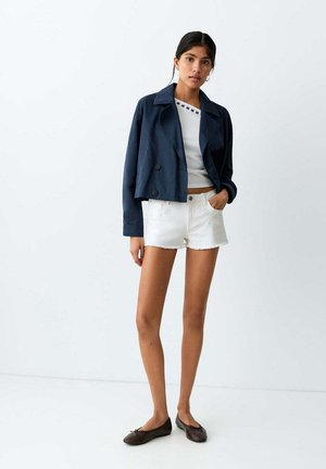 Femme portant une veste courte bleu marine, un débardeur blanc brodé, un short en jean blanc et des ballerines marron foncé, debout sur fond blanc.