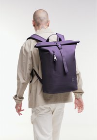 Zaino a rotolo viola con spallacci regolabili, dettagli con zip e superficie testurizzata, su uno sfondo neutro.