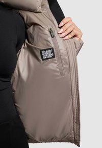 Padded jacket w kolorze taupe, z czarnym wnętrzem i kieszenią na zamek. Zawiera naszywkę z logo oraz widoczne detale szwów.