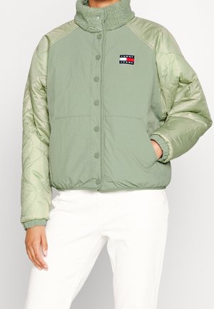 Person iført en salviegrøn Tommy Jeans jakke med quiltede ærmer, trykknapper og en sherpa krave, kombineret med hvide bukser.
