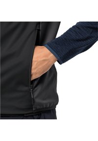 Black softshell jacket with a zippered pocket; blue textured long sleeve shirt visible at the wrist. Smooth material, fitted design.