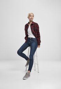 Crvena i crna karirana flanelska košulja preko bijele majice, plave uske traperice i sivi tenisice. Model sjedi na bijelom stolcu.