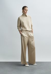 Beiger Strickrollkragenpullover kombiniert mit beigen, weit geschnittenen Satin-Hosen. Die Schuhe sind weiße, spitz zulaufende Absätze; große goldene Creolen als Detail.