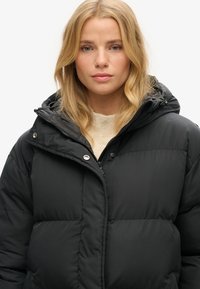 Sort puffjakke med lynlås fortil, trykknapper, stor hætte og quiltet struktur. Bæres over en lys beige sweater.