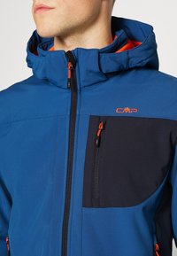 Kék softshell dzseki kapucnival, fekete díszítéssel, narancssárga cipzárfogókkal és cipzáras mellzsebbel. CMP logó a mellkason.