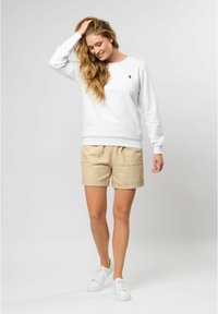 Balta sporta krekla ar apaļu kakla izgriezumu un melnu logo, apvienota ar beigu šortiem, kuriem ir divas priekšējās kabatas un ruļļoti apakšmalas. Baltas sporta apavi.