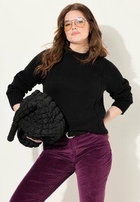 Zwarte gebreide trui met geribde manchetten, gecombineerd met paarse corduroy broek. Een gewatteerde zwarte handtas wordt onder de arm gehouden.