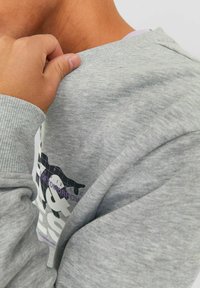 Sudadera gris hecha de un tejido suave, con puños y cuello de canalé. Muestra un diseño gráfico colorido en la parte frontal en tonos púrpura y negro.