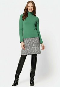 Frau in grünem Rollkragenpullover, schwarz-weiß gemustertem Rock, schwarzen Strumpfhosen und kniehohen schwarzen Lederstiefeln, stehend und lächelnd.