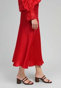 Jupe maxi en satin rouge avec un ourlet évasé, associée à des sandales à talons blocs strappées marron. Texture lisse et design fluide.