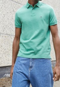 Green cotton polo shirt with a collared neckline and short sleeves, featuring a small logo. Paired with light blue denim jeans.