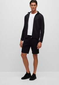 Svart zip-up jacka med huva, kombinerad med svarta shorts. Under finns en vit t-shirt. Modellen har på sig svarta sportskor och står framför en enfärgad bakgrund.