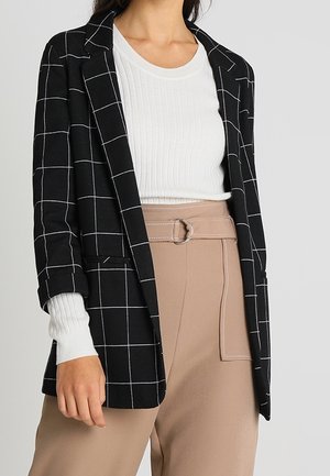 Frau trägt einen schwarzen karierten Blazer über einem weißen, gerippten, bauchfreien Oberteil und eine hoch taillierte beige Hose mit Gürtel und Ringschnalle.