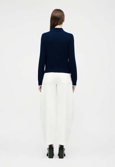 Femme aux longs cheveux bruns portant un haut à manches longues bleu marine, un pantalon blanc à jambes larges et des chaussures à talons noirs, debout de dos.