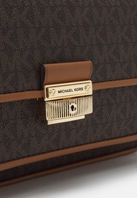 Brązowa torebka o teksturowanej powierzchni z złotym zapięciem z logo i brązowymi skórzanymi akcentami, prezentująca wzorzysty zewnętrzny wygląd.