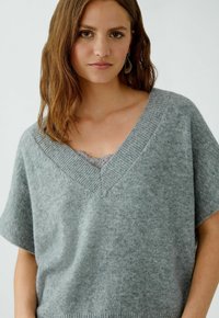 Frau mit braunem Haar, die einen grauen V-Ausschnitt-Kurzarmpullover über einem Spitzenoberteil trägt, mit hängenden Ohrringen, vor einem einfarbigen Hintergrund.