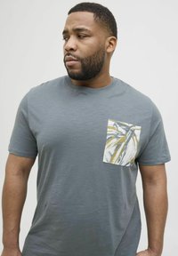 T-shirt gris à manches courtes en matériau doux. Présente une poche blanche avec un motif de feuille de palmier dans des nuances de jaune et de bleu.