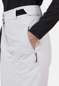 Bílé lyžařské kalhoty s texturovanou tkaninou, bočními kapsami na zip a páskem na suchý zip. Přiléhavé, s hladkým povrchem a ergonomickým designem.