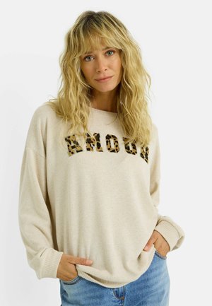 Femme blonde aux cheveux ondulés portant un pull beige avec un texte léopard "BROAD", les mains dans les poches, associé à un jean en denim bleu.