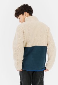 Beige fleece pullover with textured curly fabric and dark blue flat fabric panel at the bottom. Features a high collar and long sleeves.
