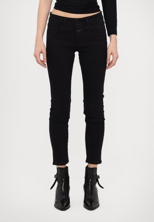Femme portant un jean noir skinny raccourci et des bottines noires à bouts pointus, debout devant un fond blanc.
