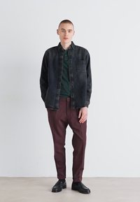 Camicia di jeans nera sovrapposta a un polo verde scuro, abbinata a pantaloni tapered borgogna e scarpe nere, su uno sfondo neutro.