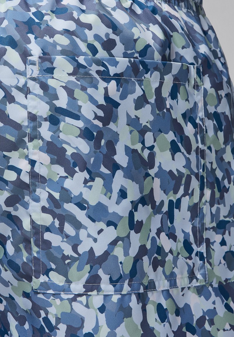Un tissu à motifs bleus et verts présente des formes abstraites et une poche cousue, avec une texture lisse et une taille élastique.