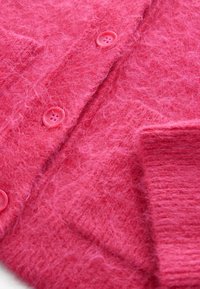 Cardigan tricotat dintr-un material roz vibrant și pufoasă, având două butoane roz și detalii cu manșete cu oarecare textură. Textură moale cu un ușor luciu.