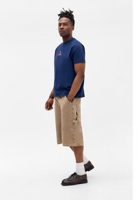 Man standing wearing navy blue t-shirt, beige knee-length shorts with side pocket, white socks, and brown leather shoes against white background.