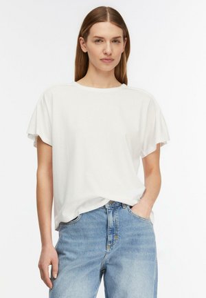 Kobieta z prostymi brązowymi włosami, ubrana w luźną białą koszulkę z krótkim rękawem i jasnoniebieskie jeansy z wysokim stanem, z jedną ręką w kieszeni.