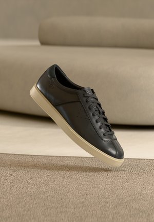 Black leather lace-up sneaker with beige sole floating above beige carpet in front of a beige couch in a minimalistic setting.