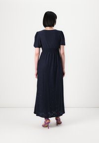 Marineblå maxi-klänning med korta puffärmar, smockad midja och detaljer i ögonbroderi. Kombinerad med lila högklackade sandaler.