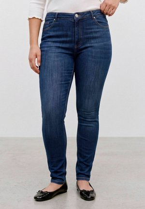 Kvinde iført mørkeblå skinny jeans og sorte balletsko, med den ene hånd hvilende ved taljen og den anden afslappet langs siden.