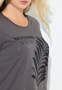 Harmaa T-paita pyöreällä kaula-aukolla, koristeltu mustalla lehtikuvioinnilla ja tekstillä "MODERN VIEW", jossa on pieniä metallinhohtoisia niittejä.