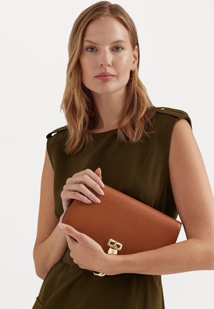 Vrouw met schouderlang blond haar, gekleed in een mouwloos olijfgroen topje, die een bruine leren clutch met gouden sluiting vasthoudt.