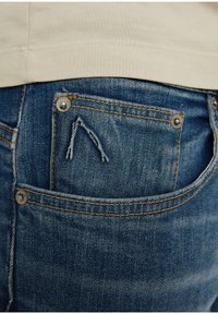 Džínové kalhoty modrého odstínu, s malou opotřebovanou zadní kapsou a stříbrným kováním. Textura je hladká s viditelným šitím.