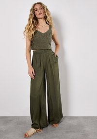 Modèle portant un top sans manches olive avec broderies florales, assorti à un pantalon olive ample et des sandales simples, posant devant un fond neutre.