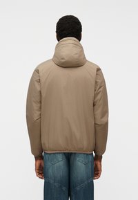 Άνδρας που φοράει μπεζ μπουφάν με κουκούλα και μπλε τζιν, στέκεται με την πλάτη προς την κάμερα μπροστά σε ένα απλό φωτεινό φόντο.