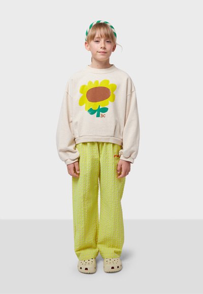 Sweatshirt in light beige with a sunflower design, paired with bright green textured wide-leg pants and beige clogs. Hair tied with a striped headband.