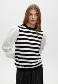 Maglione a righe in bianco e nero con maniche corte a sbuffo bianche e dettagli in oro sui bottoni delle spalle. Presenta un design aderente e orlo a coste.