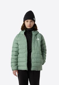 Hele roheline kapuutsiga puffer-jope, millel on ees tõmblukk, tikandiga muster ja rinnal logo, kantuna musta pooltrikoo all.