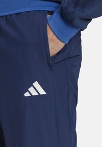 Marineblaue Sporthose mit strukturiertem Finish, featuring ein weißes gesticktes Adidas-Logo und sichtbare Seitentasche. Bünde in einem helleren Blau.