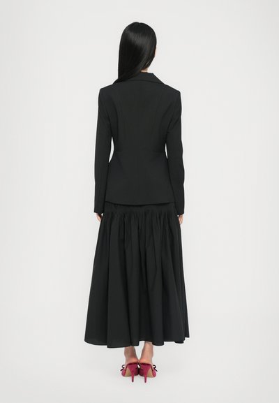 Black tailored blazer with a fitted bodice and a black maxi skirt featuring gathered waist. Pair of pink high-heeled shoes with bows.