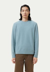 Helles blaues Sweater aus weicher Stoffqualität, Rundhalsausschnitt, lange Ärmel, lockere Passform und Bunddetails. Getragen mit braunen Shorts.