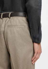 Brown beige trousers with a textured finish, featuring a dark brown belt and back pocket; paired with a dark, lightweight, buttoned shirt.