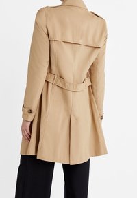 Beige trench coat with a belted waist, double-breasted buttons, and back pleat details. Made of smooth fabric, featuring long sleeves.