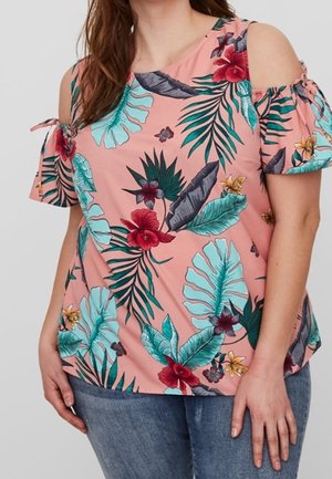 Kvinde iført lyserød bluse med bar skulder og farverigt tropisk blomstermotiv samt blå jeans, stående foran en ensfarvet baggrund.