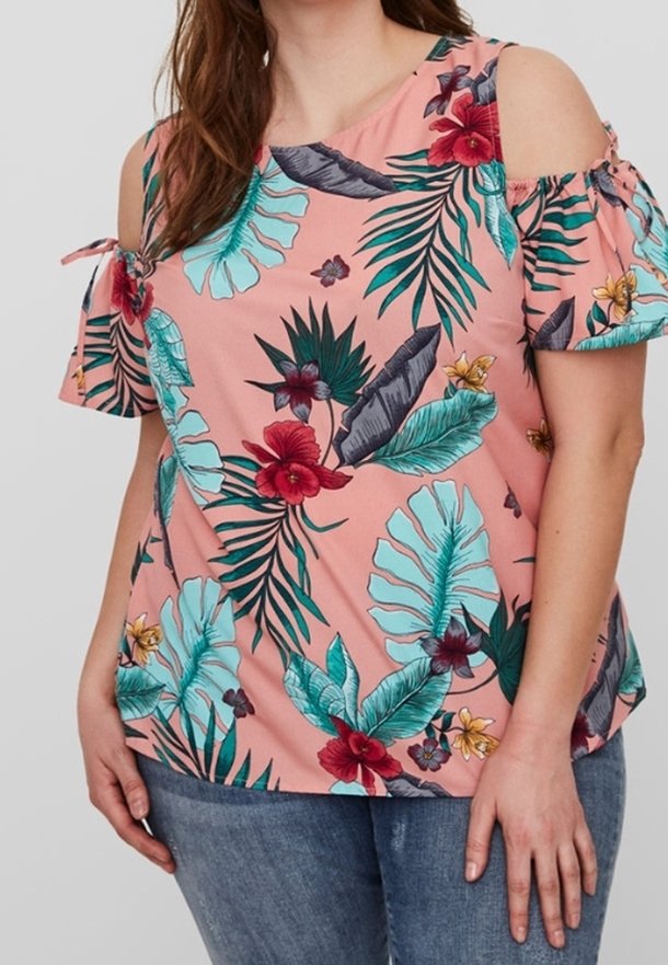 Kvinde iført lyserød bluse med bar skulder og farverigt tropisk blomstermotiv samt blå jeans, stående foran en ensfarvet baggrund.