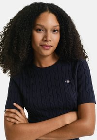 Donna con capelli ricci che indossa un maglione blu scuro a maniche corte con trecce, braccia incrociate, con espressione neutra, rivolta in avanti.