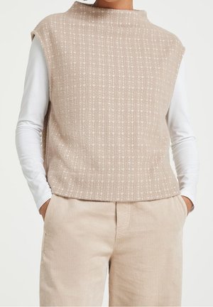 Veste sans manches beige en maille texturée portée par-dessus un haut à manches longues blanc, associée à un pantalon en velours côtelé beige, mains dans les poches.